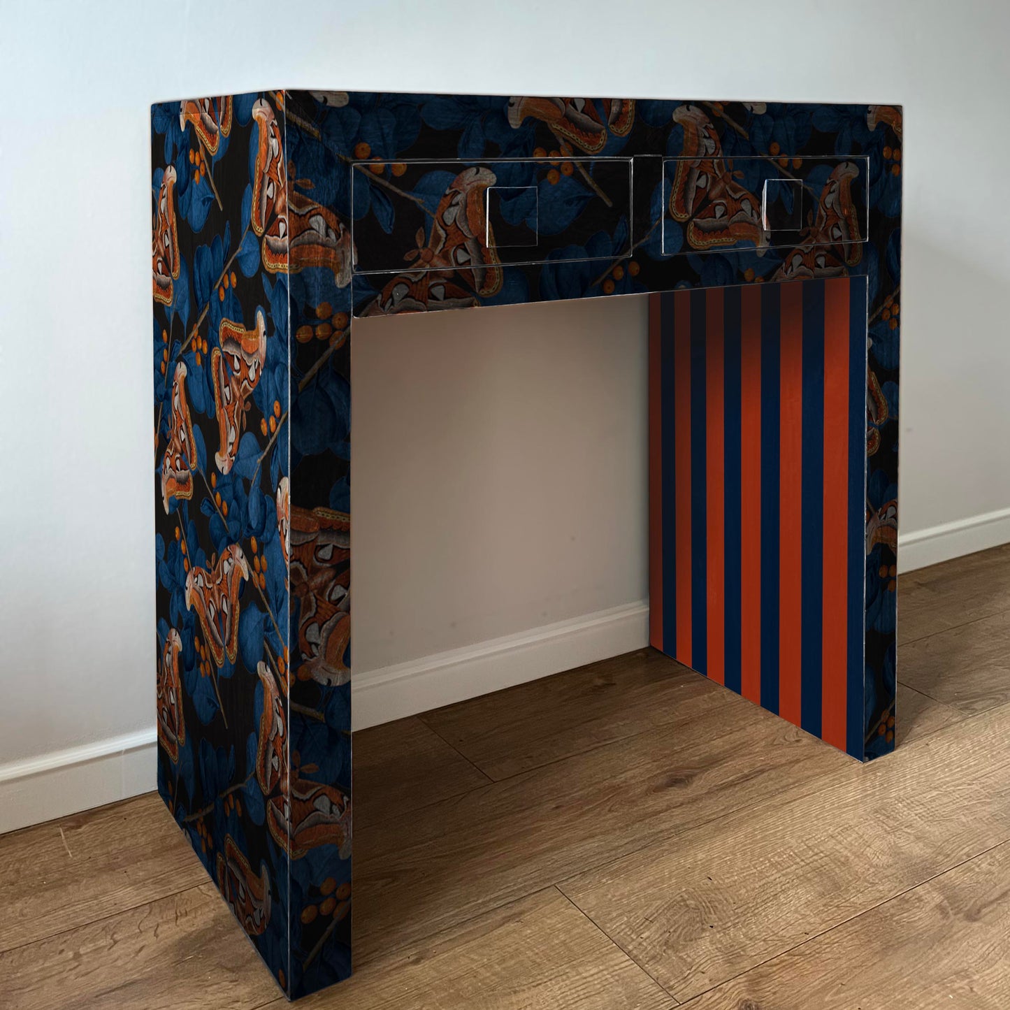 Decorative console table with moth in dark background with blue leaves and striped design against a plain wall.